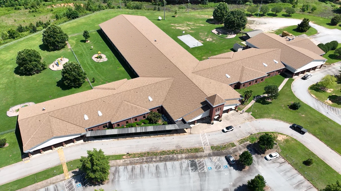 Top view of an elementary school building with brown asphalt shingle roofing, showing the layout of the roof and surrounding school grounds.