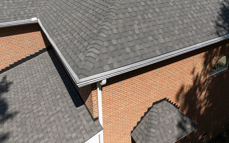Close-up view of a house with gray asphalt shingle roofing and clean, silver metal gutters installed along the roof edges, attached to a red brick exterior wall